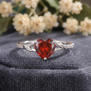 May include: A silver ring with a heart-shaped, deep red gemstone. The ring features a delicate leaf design on the sides of the gemstone and small accent stones along the band. The ring is set against a gray background.