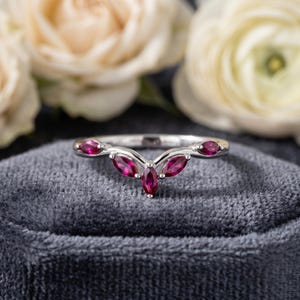 May include: A silver ring with a curved design, featuring five marquise-cut, deep red gemstones. The ring is set against a gray velvet surface, with blurred white flowers in the background.