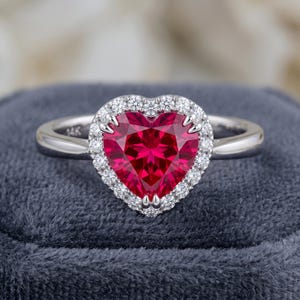 Könnte beinhalten: Ein herzförmiger Rubinring mit einem Kranz aus kleinen, klaren Diamanten. Der Rubin hat eine leuchtend rote Farbe und ist in einem silbernen Band gefasst. Der Ring wird auf einer dunkelgrauen Samtoberfläche präsentiert.