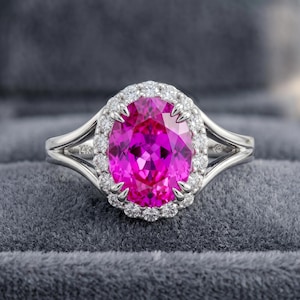 Puede incluir: Un anillo de plata con una gran gema rosa brillante de talla ovalada, rodeada por un halo de piedras más pequeñas y claras. La banda del anillo se divide en un diseño de doble caña. El anillo está sobre un fondo gris.