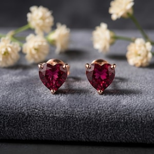 May include: A pair of heart-shaped stud earrings. Each earring features a deep red gemstone set in a gold-colored metal. The gemstones are cut to resemble a heart. The earrings are displayed on a gray surface.