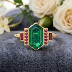 May include: A gold ring with a central emerald-cut green gemstone, framed by smaller red gemstones. The ring has a geometric design and a gold band. The ring is displayed on a grey surface, with flowers in the background.