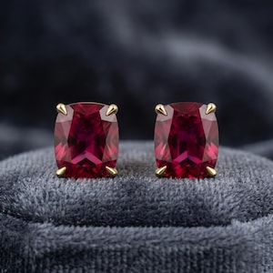 Può includere: Un paio di orecchini a bottone quadrati con pietre preziose rosso scuro. Ogni orecchino presenta una montatura a griffe color oro che tiene la pietra sfaccettata a taglio cuscino. Gli orecchini sono esposti su una superficie di velluto grigio scuro.
