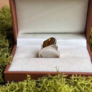 May include: A silver ring featuring a single, rectangular, polished amber stone set at an angle. The ring is displayed inside a brown, rectangular jewellery box with a white interior lining, positioned to showcase the metallic band and the stone.