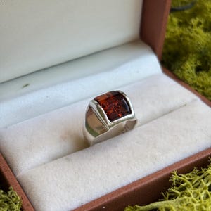 May include: A sterling silver ring featuring a rectangular, polished amber gemstone set in a smooth, wide band. The metal band is stamped with 925, indicating its silver content, and the amber displays a deep, translucent reddish-brown hue.