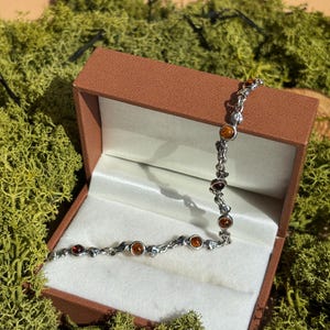 May include: A silver bracelet with amber stones, presented in a brown jewellery box lined with white satin. The bracelet is displayed on a bed of green moss.