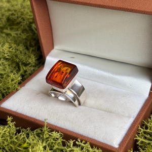 May include: A silver ring featuring a rectangular, polished amber stone set in a smooth metal bezel. The amber is a deep, translucent orange colour with visible internal inclusions. The ring band is wide, polished silver with a split design.