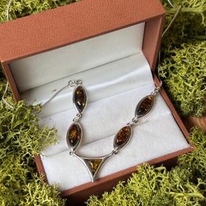 May include: A silver necklace featuring four marquise-shaped amber stones and one triangular amber stone set in metal links. The necklace is displayed inside an open brown jewelry box with a white interior lining, resting on green moss.