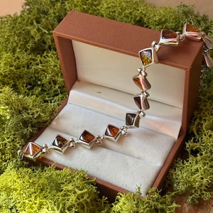May include: A silver link bracelet featuring multiple square-shaped amber stones in varying shades of orange, yellow, and brown. The bracelet is displayed inside an open brown jewellery box with a white interior lining.