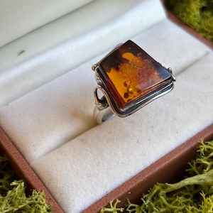 May include: A silver ring featuring a large, trapezoidal, polished amber gemstone with deep orange and brown hues. The setting includes intricate silver wire detailing along the sides, securing the stone within a metallic frame.