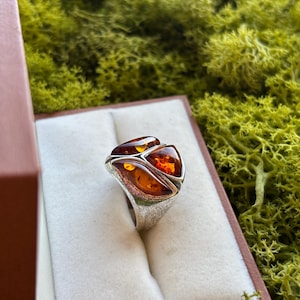 May include: A silver ring featuring three triangular, polished amber stones set in a textured metal band. The ring is displayed inside a small, open box with a white velvet interior, showcasing the warm, translucent orange hues of the amber.