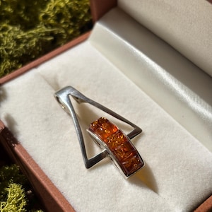 Può includere: Ciondolo in argento con cornice triangolare e ambra rettangolare. Il ciondolo è presentato in un cofanetto marrone con fodera in raso bianco. L'ambra ha un colore caldo, arancione-marrone.