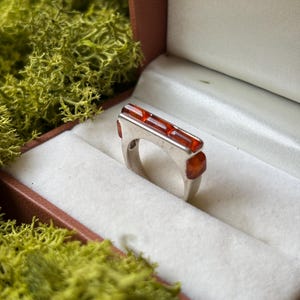 May include: A silver ring featuring a rectangular top set with three smooth, translucent amber stones. The ring has a polished metallic band and is displayed in a white-lined box, showcasing a clean, geometric design with warm orange tones.