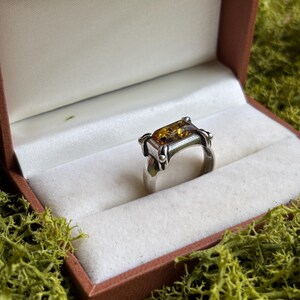 Op de afbeelding: Een zilveren ring met een rechthoekige, doorschijnende barnstenen steen in een klauwzetting. De ring heeft een gepolijste metalen band en wordt gepresenteerd in een bruine doos met een witte voering, wat de warme goudbruine tinten benadrukt.