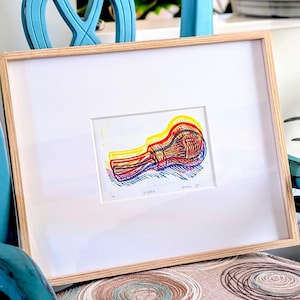 May include: A framed art print featuring a colorful abstract design in red, yellow, and blue. The artwork is displayed on a white mat within a natural wood frame. The print is resting on a chair with a patterned seat and a turquoise blue frame.