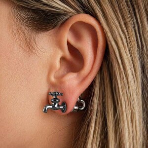 May include: A close-up of a person's ear with a silver faucet earring. The earring is a detailed miniature replica of a water tap. The earring is worn on the earlobe. The background is blurred.