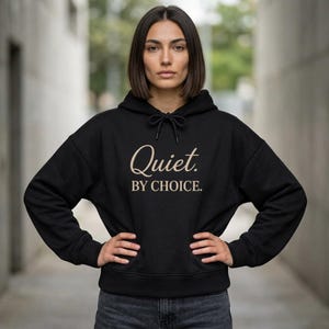 May include: Black hoodie with the text "Quiet. BY CHOICE." in a cursive font. The hoodie has a drawstring and a kangaroo pocket. The model is wearing dark wash jeans.
