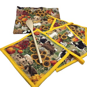 May include: A set of quilted farm-themed kitchen accessories, including pot holders and a trivet. The fabric features cows, sunflowers, pumpkins, and a red barn. The pot holders have yellow trim and loops. A wooden spoon rests on the trivet.