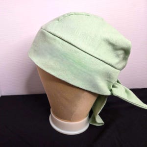 May include: A light green head wrap, likely made of cotton, is displayed on a mannequin head. The head wrap is tied at the back, with the ends hanging down. The fabric has a slightly textured appearance.
