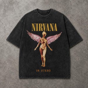 Peut inclure: Un t-shirt noir délavé à l'acide avec un motif représentant une silhouette ailée à la peau dorée et aux ailes roses. Le texte NIRVANA est imprimé en doré au-dessus de la silhouette, et le texte IN UTERO est imprimé en doré en dessous.