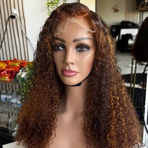 Pode incluir: Uma cabe&ccedil;a de manequim exibindo uma peruca castanha encaracolada com reflexos. A peruca apresenta uma frente de renda e &eacute; estilizada com cachos volumosos. A cor do cabelo transita de um castanho mais escuro nas ra&iacute;zes para reflexos castanhos dourados mais claros.