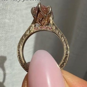 May include: A gold ring featuring a large, faceted, light pink center stone held in a floral-shaped setting. The band is decorated with intricate leaf-like engravings and a row of small pink accent stones along the upper shank near the setting.