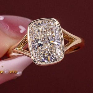 May include: A gold ring featuring a large, rectangular cushion-cut center stone with rounded corners, secured in a bezel setting. The band has a split-shank design that tapers toward the center stone, creating a polished and metallic appearance.