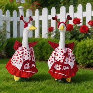 May include: Two white goose statues with yellow beaks and feet, adorned with red heart-shaped headbands and wings. They wear white dresses patterned with red hearts and red ruffled skirts, standing on green grass in front of a white picket fence.