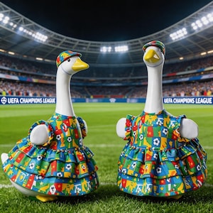 May include: Two ceramic goose figurines wearing colorful soccer-themed dresses and caps, set against a stadium backdrop. The dresses feature a vibrant pattern of soccer balls, flags, and geometric shapes in blue, green, red, and yellow.