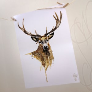 May include: A watercolour print of a stag with large antlers. The artwork uses a brown and tan colour palette with a splattered effect, creating a textured appearance. The print is suspended from a string with wooden pegs.