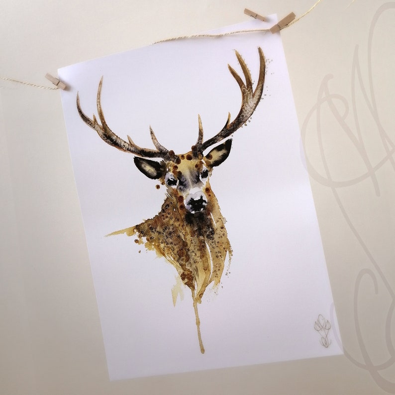 May include: A watercolour print of a stag with large antlers. The artwork uses a brown and tan colour palette with a splattered effect, creating a textured appearance. The print is suspended from a string with wooden pegs.