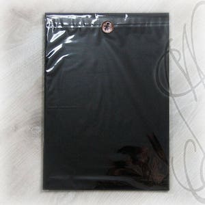 May include: A black, rectangular package sealed in clear plastic. A decorative button with a floral design secures the top. The package is likely for shipping or presentation of an item.