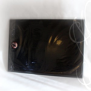 Puede incluir: Un archivador negro brillante con cierre de bot&oacute;n. El archivador est&aacute; sellado en pl&aacute;stico transparente. Se ve un peque&ntilde;o bot&oacute;n redondo con un dise&ntilde;o floral en la portada. El archivador parece nuevo y sin usar.