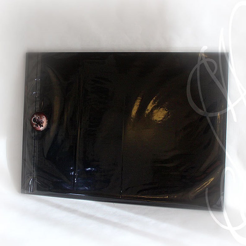 Puede incluir: Un archivador negro brillante con cierre de bot&oacute;n. El archivador est&aacute; sellado en pl&aacute;stico transparente. Se ve un peque&ntilde;o bot&oacute;n redondo con un dise&ntilde;o floral en la portada. El archivador parece nuevo y sin usar.