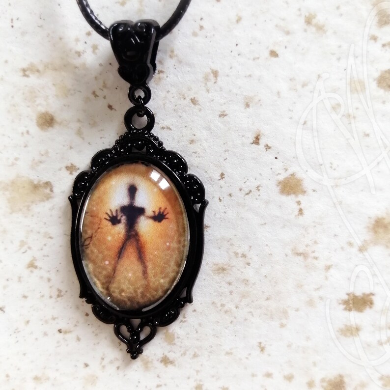 Puede incluir: Collar con colgante ovalado negro. El colgante presenta una figura silueta con los brazos extendidos sobre un fondo naranja y amarillo. El colgante est&aacute; enmarcado en un marco decorativo de metal negro y cuelga de un cord&oacute;n negro.