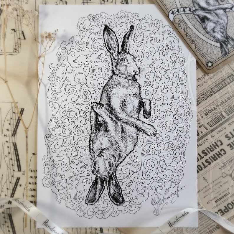 K&ouml;nnte beinhalten: Schwarz-Wei&szlig;-Tuschezeichnung von zwei Hasen, einer aufrecht und einer umgekehrt, in einem wirbelnden, ornamentalem Design. Das Kunstwerk ist mit "Martine" signiert und enth&auml;lt das Wort "Handmade" auf einem Band. Eine kleine dekorative Schachtel mit einem Hasenbild ist sichtbar.