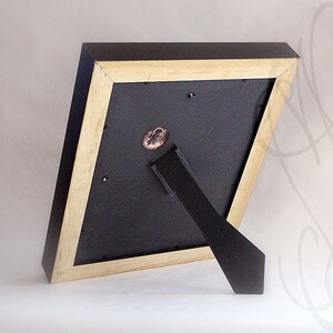 K&ouml;nnte beinhalten: Ein schwarzer und naturbelassener Holzbilderrahmen, von hinten betrachtet, mit schwarzer R&uuml;ckwand und schwarzem Staffeleist&auml;nder. Der Rahmen hat eine geometrische Form und ein kleines, rundes Emblem auf der R&uuml;ckseite.
