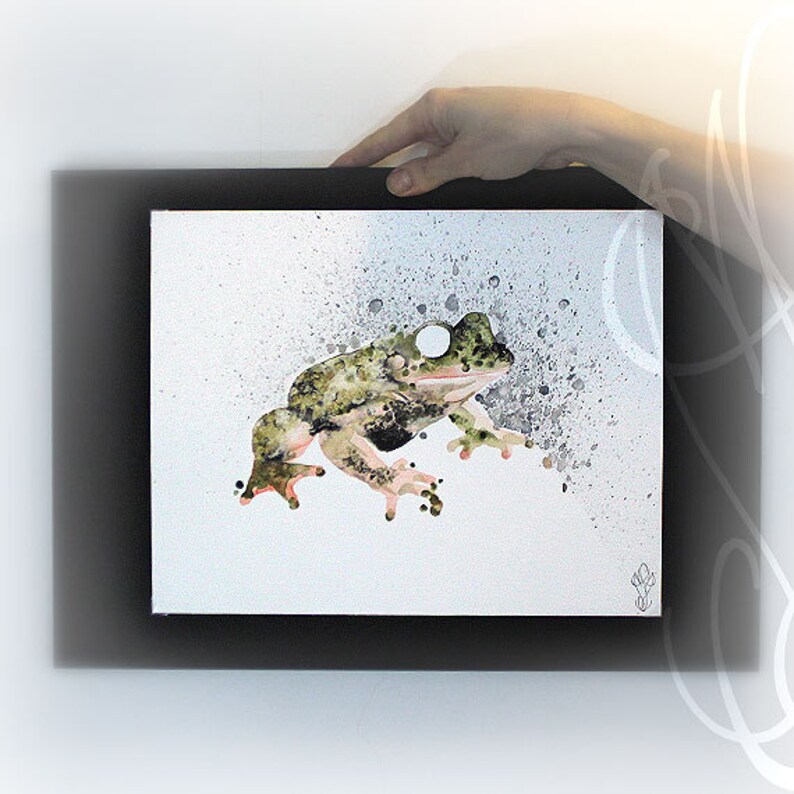 Puede incluir: Una pintura de acuarela de una rana en tonos verdes, negros y marrones, con un fondo blanco y un marco negro. La rana se representa de perfil con un patr&oacute;n moteado. Efectos de salpicaduras rodean la rana.