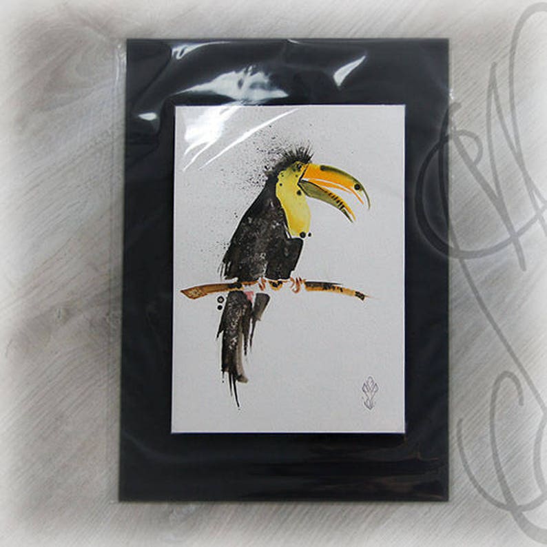 May include: A watercolor painting of a toucan bird perched on a branch. The bird has a large yellow and orange beak, black and yellow feathers, and is set against a white background. The artwork is framed in a black frame.