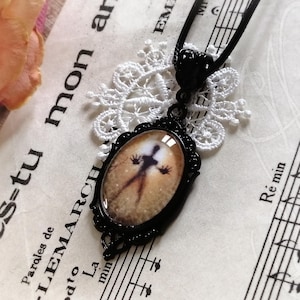 Puede incluir: Collar con colgante ovalado negro con un dise&ntilde;o de silueta espeluznante. El colgante tiene un marco de filigrana negro y cuelga de un cord&oacute;n negro. El fondo incluye partituras y un tapete de encaje blanco.