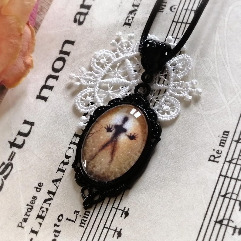 Puede incluir: Collar con colgante ovalado negro con un dise&ntilde;o de silueta espeluznante. El colgante tiene un marco de filigrana negro y cuelga de un cord&oacute;n negro. El fondo incluye partituras y un tapete de encaje blanco.
