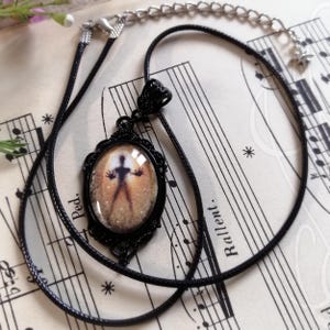 Puede incluir: Collar con colgante negro. El colgante ovalado presenta una silueta sobre un fondo dorado, enmarcada en un marco decorativo de metal negro. El collar tiene un cord&oacute;n negro y un extensor de cadena. El dise&ntilde;o del colgante evoca una est&eacute;tica vintage.