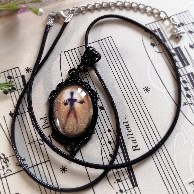 Puede incluir: Collar con colgante negro. El colgante ovalado presenta una silueta sobre un fondo dorado, enmarcada en un marco decorativo de metal negro. El collar tiene un cord&oacute;n negro y un extensor de cadena. El dise&ntilde;o del colgante evoca una est&eacute;tica vintage.