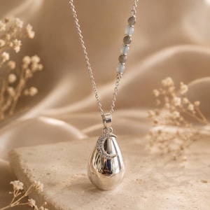Pode incluir: Um colar de prata com um pingente em forma de gota adornado com uma pequena lua crescente incrustada com pedras claras. A corrente inclui uma secção de contas alternadas em cinzento e translúcido, criando um acessório delicado.