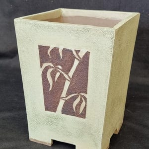 May include: A square, light cream-coloured planter with a textured, matte finish. The front panel features a recessed, dark brown rectangular inlay decorated with a carved bamboo stalk design. The planter stands on four small, integrated feet.