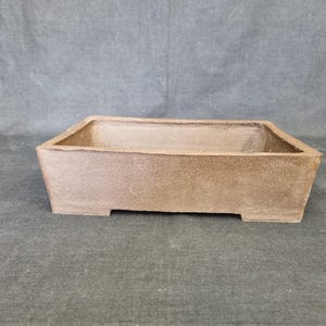 May include: A rectangular, brown bonsai pot with a textured surface. The pot has a slightly trapezoidal shape with a flat base and small feet. The interior is a lighter shade of brown.