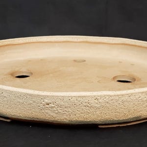 May include: An oval-shaped bonsai pot in a light brown hue, featuring a textured surface. It has two drainage holes and a raised rim. The design is simple and functional, suitable for bonsai cultivation.