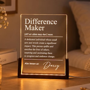 Könnte beinhalten: Eine rechteckige, klare Acrylplatte mit einem dunklen Holzsockel, die von unten beleuchtet wird. Die Platte zeigt einen weißen Text, der den Begriff Difference Maker definiert, gefolgt von dem Namen Darcy in einer leuchtenden Schreibschrift.