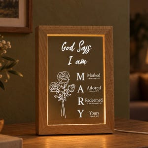 Può includere: Una scatola luminosa rettangolare con cornice in legno che presenta il testo God Says I am MARY e un'illustrazione di rose. Le lettere M, A, R, Y corrispondono alle parole Marked, Adored, Redeemed e Yours, seguite da riferimenti biblici.