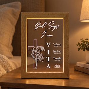 May include: A rectangular light-up sign with a wooden frame featuring the text God Says I am VITA, with V for Valued, I for Invited, T for Taught, and A for Alive, accompanied by Bible verse references and a line-art cross with floral accents.